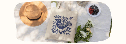 Tote bag motif oiseau bleu sur nappe, près d'un chapeau, fleurs et fruits, style streetwear écolo